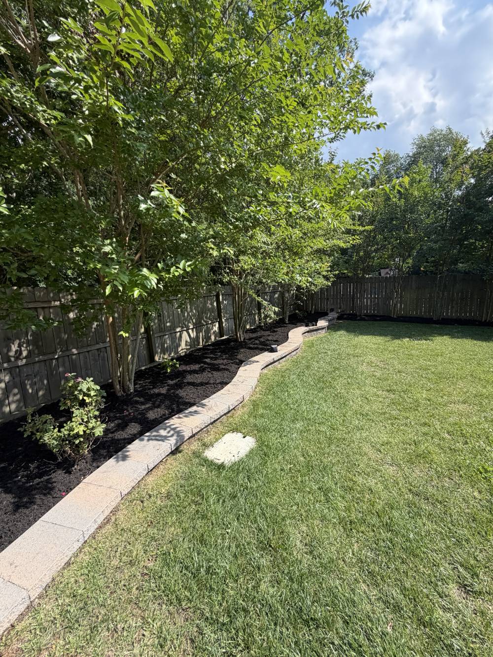 Backyard bed along a wood fence with fresh black mulch behind a curved stone border